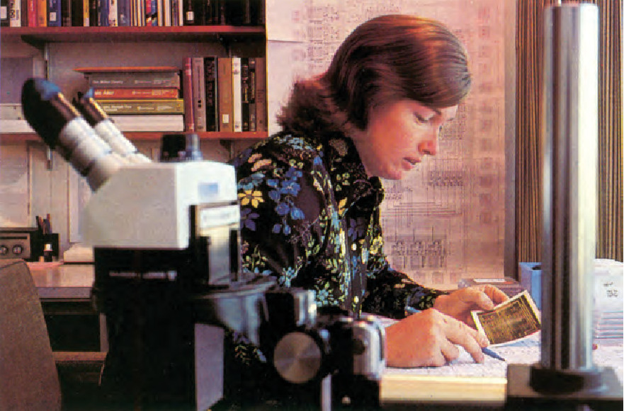 Lynn Conway trabajando mirando un papel comparando algo ocn una ficha en su otra mano. Al fondo se ve una estanteria con libros y en primer plano a la izquierda un microscopio.