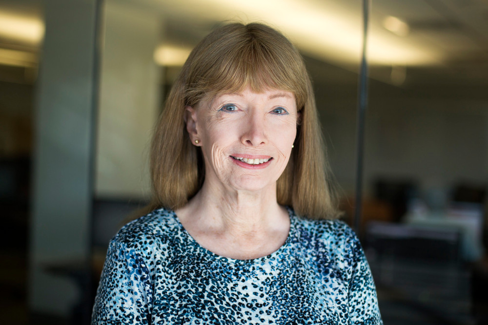 Lynn Conway mirando a cámara sonriente con una melena castaña larga, ojos azules, pendientes dorados. Fondo desenfocado en tonos dorados.