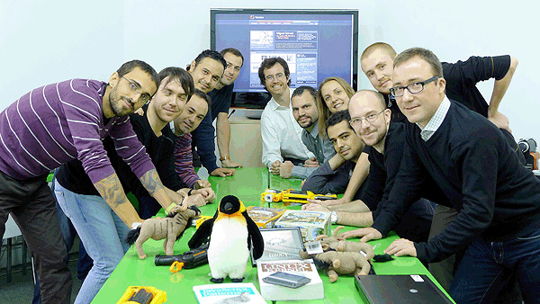 Foto del equipo de Kaleidos en 2011. 11 hombres y una mujer jóvenes posan mirando a cámara apoyados en una mesa en el medio de la cual está Tux, la mascota de Linux.