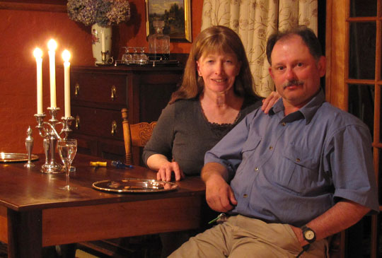 Lynn Conway mirando a cámara con una mano sobre el hombro de su marido, ambos sentados en la mesa de un comedor en la que hay un candelabro con velas encendido