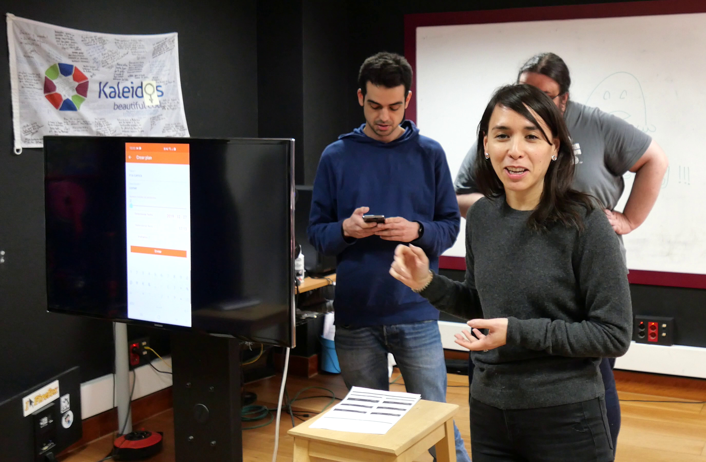 En primer plano se ve a Marina mirando a cámara y a su izquierda una pantalla donde se muestra una app en el móvil. Detrás se ve a JuanFran mirando su móvil y parte de la cabeza de Alonso tapada por Marina ;)