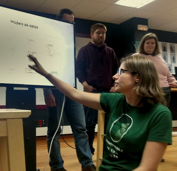 Foto de la presentación del proyecto en la oficina de Kaleidos donde se ve a Maria señalando algo en la pantalla. Lleva una camiseta verde de 'Programar es una Mierda' 
